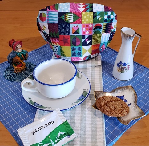 A tea tableau with a festive air featuring a cozy that looks like gift wrap, a leaf-shaped dish holding a large gingersnap cookie, a white, blue and green cup and saucer, a tiny flowered urn matching the leaf dish, and a detailed figurine of a pheasant dressed in an old-fashioned plaid cape and bonnet and bearing a basket of gifts. The tea is Yuletide Toddy from Adagio Teas.
