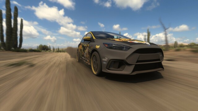 2017 Ford Focus RS with a grey wrap and Cheetos-branded livery shot offset from the front with a fisheye lens driving on a dirt road. Plumes of dust and gravel come from all four wheels and cactus and rocks line the road. The sky is bright with fluffy clouds.