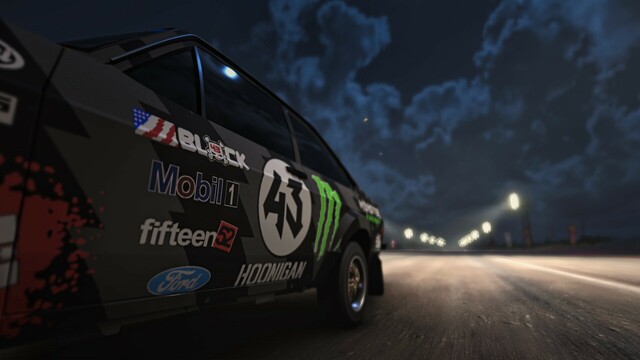 Close-up of 1977 Ford Escort RS1800 shot from ground level near the front quarter panel along the body of the car. The livery features Ken Block, Hoonigan, Ford, Mobil1, Fifteen52, and Number 43 badging. Lights line the drag strip and the moon illuminates scattered clouds in the sky.