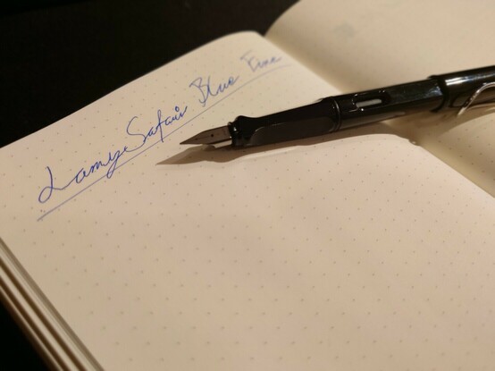 A picture of an bullet journal with blue text that says Lamy Safari Blue Fine.
A Lamy Safari pen lies next to the text.
