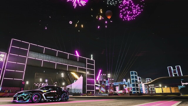Distance shot of the driver side of a black and gold 2020 Toyota GR Supra reflecting magenta, cyan, and yellow neon slights. The horizon festival stage and concert lasers are in focus in the distance. Gold and magenta fireworks fill the sky and a single hot air balloon floats into the distance.