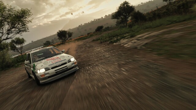 Distance shot taken from the front of a 1992 Escort RS Cosworth in a retro green, gray, and white Fujifilm livery as it drifts at full wheel lock around a dirt corner. Behind the car, the sun is nearing the horizon but obscured by clouds. Green trees and orange flowers fill the fields.