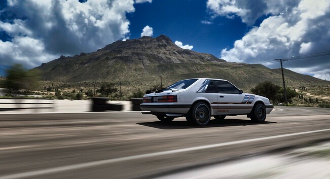 Moving shot of a white 1986 Ford Mustang SVO with chrome stripes moving from left to right in the frame on a paved road throughs sand dunes. Blue skies with thunderheads loom in the distance and a giant dormant volcano fills the horizon.