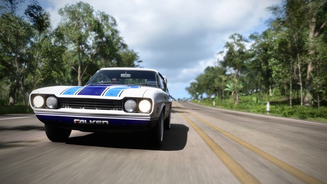 Close-up shot of 1973 Ford Capri RS3100 taken from the front. The road and trees blur with speed as the sun reflects off the dark blue and white livery. Three stripes in lighter shades of blue line the hood and the sun reflects off the chrome bumper.