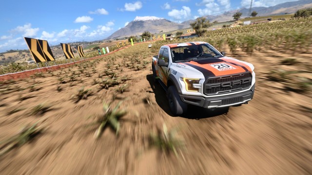 2017 Ford F-150 Raptor with a silver, black, and orange striped livery driving through an agave field with another truck in the distance. The speed and fish eye lens create extreme blur.