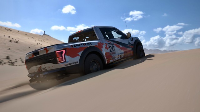 Shot from the rear passenger side of a 2017 Ford F-150 Raptor with a silver, black, and orange striped livery climbing a sand dune. Sand reaches the center of the wheels and the truck leaves deep ruts as it digs through.