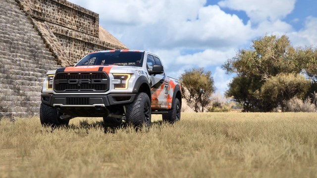 Shot from the front driver side of a 2017 Ford F-150 Raptor with a silver, black, and orange striped livery parked near a historic stone temple. Trees line the horizon and clouds billow in the sky.