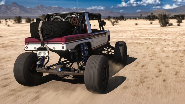 Shot from behind of a 2017 Ford #25 “Brocky” Ultra4 Bronco RTR with large offroad tire, lifted suspension, a full roll cage, light bars, and a large spare strapped to the back on the beach. The body is painted in brick red with a large white cutout and black pin striping. The truck is moving at speed through the desert. Sand kicks up off the tires and sparse brush blurs past with a village blurred in the distance. Mountains line the horizon.