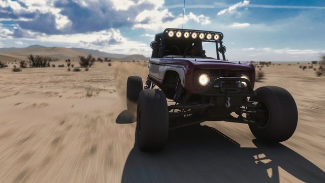 Close-up from the front of a 2017 Ford #25 â€œBrockyâ€� Ultra4 Bronco RTR with large offroad tire, lifted suspension, a full roll cage, light bars, and a large spare strapped to the back on the beach. The body is painted in brick red with a large white cutout and black pin striping. The truck is moving at speed through the desert. Sand kicks up off the tires and sparse brush blurs due to speed. Fluffy white clouds obscure the sun and fill the sky, allowing peeks of blue and reflections off the truck's shiny surface.