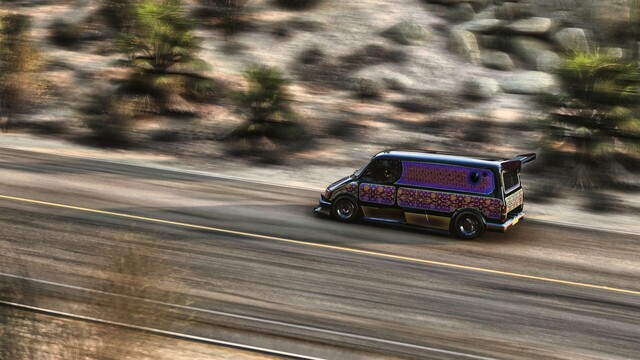 Distance shot from the drivers of a 1994 Ford Supervan 4 in a patterned black, orange, purple, and gold livery reminiscent of an ornate rug.  Cacti and brush line the road, blurred by the speed of the shot.