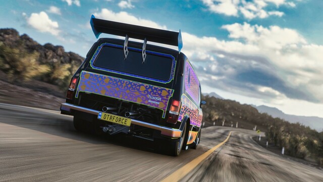 Close up shot from the passenger's side rear of a 1994 Ford Supervan 4 in a patterned black, orange, purple, and gold livery reminiscent of an ornate rug.  The van is driving down a hill, headed toward a hairpin turn. Rocks and brush blur past while large white clouds break up a deep blue sky.