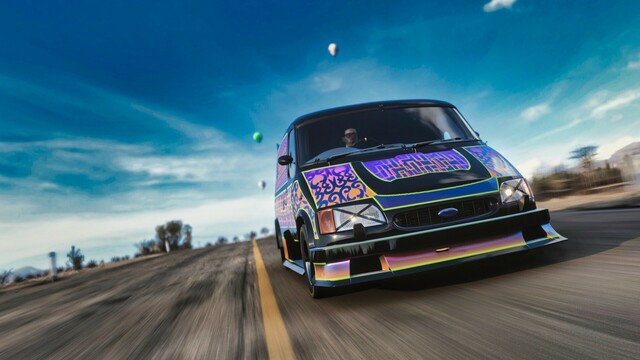 Close up shot from the front of a 1994 Ford Supervan 4 in a patterned black, orange, purple, and gold livery reminiscent of an ornate rug. The van is cresting a hill and the wispy clouds in the sky form a crown, moving away from the van in all directions. The driver focuses on the road intensely. He is wearing a red clown nose.
