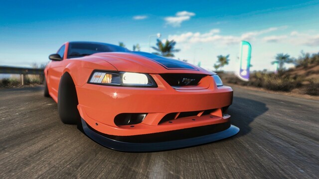 Close-up from the front of an orange and black 2000 Ford SVT Cobra R drifting with wheels at full lock. Palm trees dot the background as the rear bumper skirts next to the guard rail and the cracked asphalt blurs underneath the front bumper.