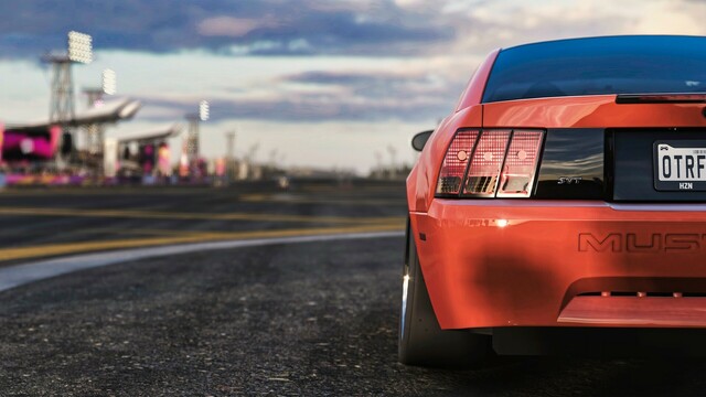 Close-up of the rear driver's side of an orange and black 2000 Ford SVT Cobra R parked on a drag strip. The wheels are cambered out and the sun reflects sharply off the rim and bumper. Festival grandstands line the drag strip.
