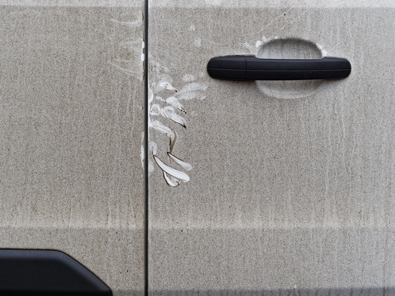 Part of the back door of a very grubby white van. The whole surface is covered with tightly packed specks of black dirt. At the edges of the door there are marks where hands have disturbed the dirt revealing the white surface beneath.
