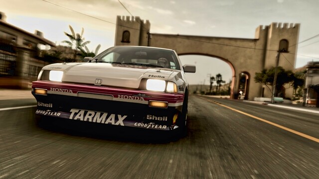 Shot from the front of a 1984 Honda CRX Mugen edition racing through a rural Mexican village. A stone archway is just behind the car and clouds fill the horizon, threatening rain.