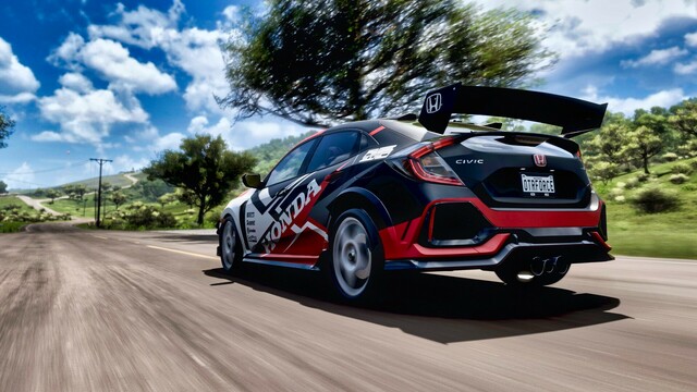 Shot from the driver's side rear of a 2018 Honda Civic Type R with an aftermarket spoiler and Team Honda red, black, white, and grey geometric livery moving at speed down a country road. Tree's blur past and green hills, blue skies, and fluffy clouds line the area.