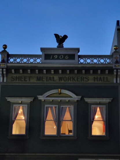 A photograph taken at twilight of the top of a historical building, windows glowing with gentle light, ''sheet metal workers hall' carved into the top, with a stone marked '1906' topped with an eagle.