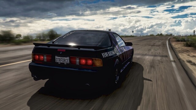 Close up capture with wide angle lens from right-hand rear of a 1990 Mazda Savannah RX-7 in a gloss black and metallic purple livery featuring stripes running diagonally along the length of the vehicle. White lettering on the rear quarter panels reads "Neon Racing" and has a smiley face with x's for eyes. The car is moving down a stretch of degrading rural desert road with storm clouds dotting the horizon. Sun glints off the rear glass.