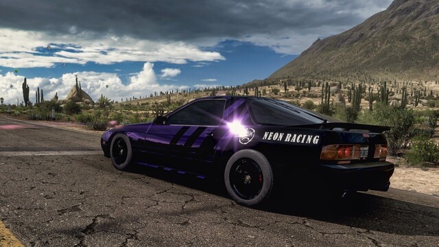 Still capture with wide angle lens from left-hand rear of a 1990 Mazda Savannah RX-7 in a gloss black and metallic purple livery featuring stripes running diagonally along the length of the vehicle. White lettering on the rear quarter panels reads "Neon Racing" and has a smiley face with x's for eyes. The car is parked on a stretch of degrading rural desert road with storm clouds dotting the horizon. Sun glints off the rear glass. Cactuses and low shrubs dot the surrounding landscape before meeting with the base of a large dormant volcano which rises up out of frame.