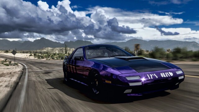 Close up capture from front of a 1990 Mazda Savannah RX-7 in a gloss black and metallic purple livery featuring stripes running diagonally along the length of the vehicle. White lettering on the front bumper reads "It's okay" with a heart. The car is moving down a stretch of degrading rural desert road with storm clouds dotting the horizon. Sun glints off the front bumper as cacti, street signs, and rocks blur past.