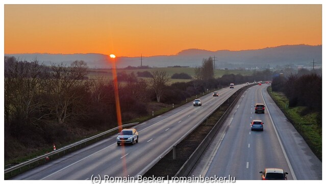 A motorway in front of a sunset over hills. The sky is red, and the sun makes a flaire.