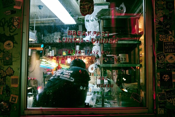 A window into a restaurant kitchen, the frame is covered in various stickers. A chef has their back to us, cleaning on the counter, resting. There's red-violent reflections in the window glass and on the frame, contrasted with the greenish light from the kitchen.