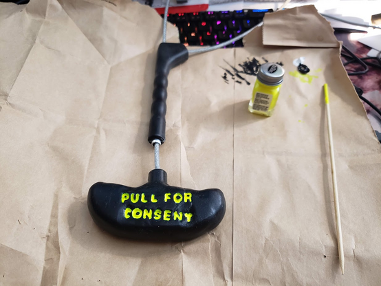 Pig snare on desk with painting supplies.  Handle is molded in black with vibrant yellow lettering reading "Pull For Consent".