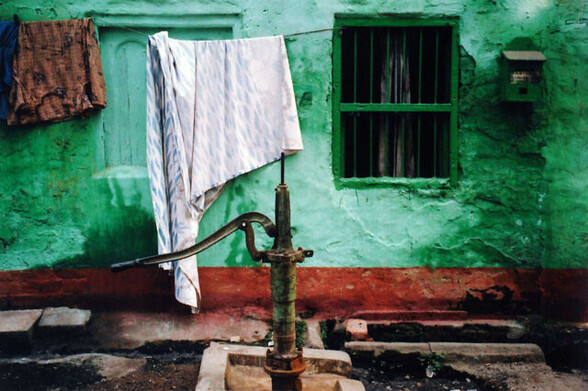 a scan of a color photo of a town pump in India. there is laundry in the background. the wall is painted a striking green. the water pump is a little rusty.