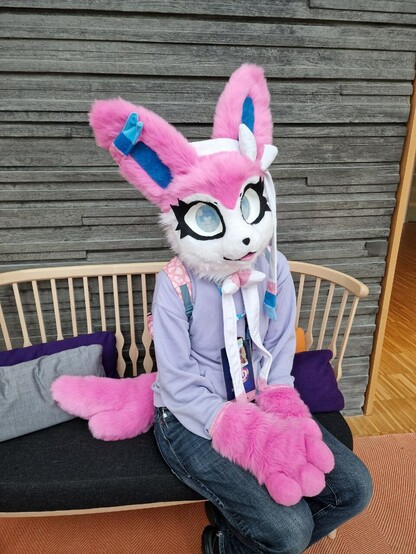 Photo of me wearing a sylveon partial fursuit, sitting on a bench against a grey wall. The fursuit is pink and white and blue and fluffy and very cute. Besides the fursuit I'm wearing a light purple hoodie and dark jeans.