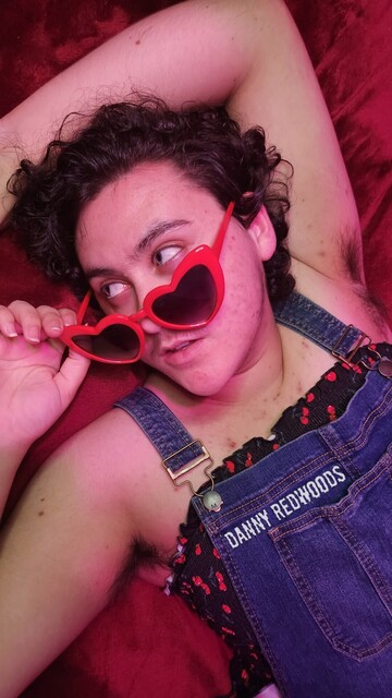 A young white trans man with curly brown hair laying on red velvet with one arm laying above his head. He's looking off to the side, peaking over the red, heart-shaped sunglasses he's wearing that he's pulling down with one hand. His lips are parted slightly, showing a hint of his front teeth and tongue. His expression is difficult to read, but he looks interested in whatever he's looking at. He's wearing blue denim overalls over a ruffled black tube top patterned with red cherries. His armpits are visible and hairy. He has a slight mustache and a variety of moles and acne scars across his skin. His eyes are a sort of blue green grey.
