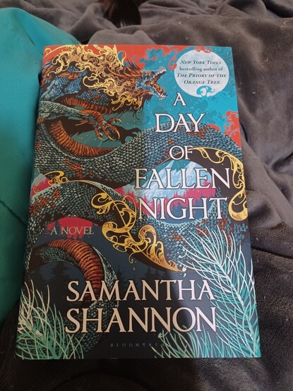 A hardcover copy of A Day of Fallen Night, by Samantha Shannon. It's the prequel to Priory of the Orange Tree. Cover features a blue, red, and gold Asian dragon against a blue and red background.