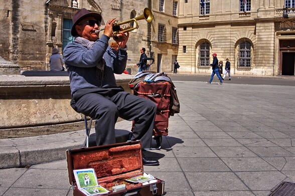 Trumpet player seated on a folding chair in an old town square. He has his instrument case open on the ground for donations and CDs for sale. He is wearing a brimmed hat and dark blue shirt and pants; the buildings in the background are shades of terracotta.