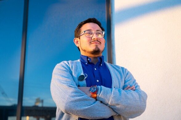 Andrew Gonzales in a blue button down sweater over a dark blue button down, and wearing horn-rimmed glases. He stands with his arms crossed proudly.