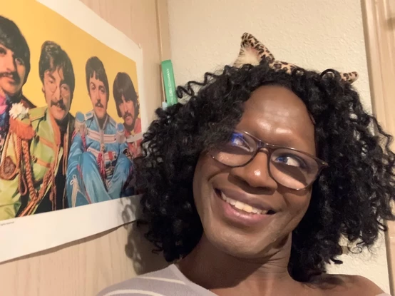 Me with curly hair and wearing glasses with the leopard print cat ears I'm looking to my right to a poster from the Beatles' *Sergeant Pepper's* album