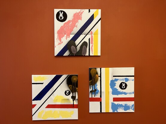 Picture of 3 abstract paintings mounted on an orange wall. Straight black lines, primary colour lines and spray painted bunny in centre of all three