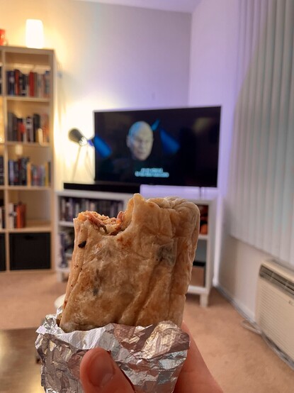 A burrito with a bit out of it, held up in front of a TV with Patrick Stewart on screen