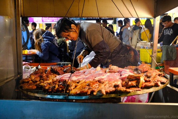 color photo of a woman making BBQ of varies meat at the streetside food stall with groups of people walking pass in the background