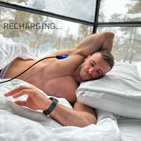 A shirtless male android powers up in bed with its power cord coming out of its armpit traveling to an off-camera power outlet as the droid checks its power level on its watch