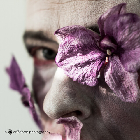color photography in 1:1 format: female self-portrait (headshot). the woman whose face is depicted in a cropped three-quarter front view has pale make-up, her eyes are emphasized darkly with a reddish rim. her mouth is covered by a dry orchid flower (partially visible in the photo) in this last of three images in the series. instead of her eye most turned towards the viewer she has another light cyclamen dry orchid (cattleia) flower. the other eye continues to look at the viewer. there is not much to see of the strict mother, who looks with a punishing look at a child who has acted contrary to her orders. but the uneasiness caused by the one eye directed at the viewer remains. you are never safe...