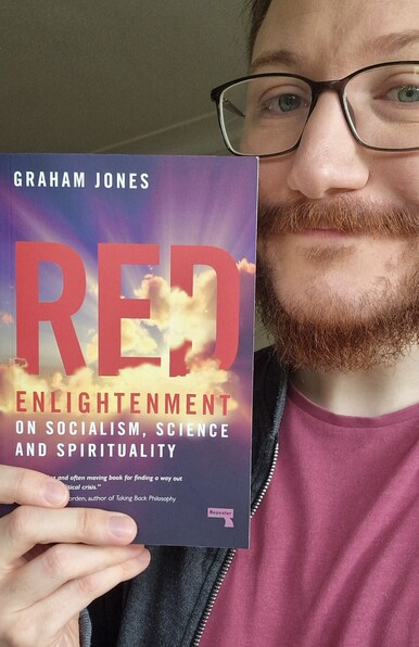 Picture of book Red Enlightenment: On Socialism, Science and Spirituality, cover is purple sky and yellow clouds with red and white writing. Book is held by a smiling man with glasses and a beard
