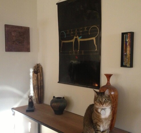 My cat sits among other objets d'art: 3 vases (turned wood, clay, hammered metal) and an Egyptian-style cat figurine on a shelf, below a dramatic black and gold poster for the Black Cat Cafe, formerly of Ottawa.

A small 3D acrylic wall piece and a small glass and mixed media piece flank the poster on the wall.