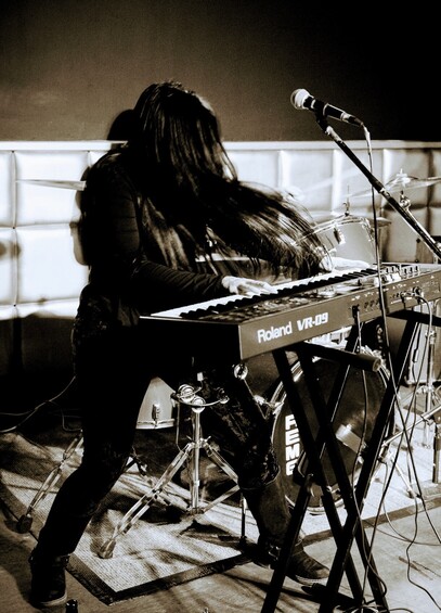A woman wearing black with long black hair swishing in front of her face plays a Roland keyboard on a stand, with a drumkit behind. There is a microphone stand in front of her.