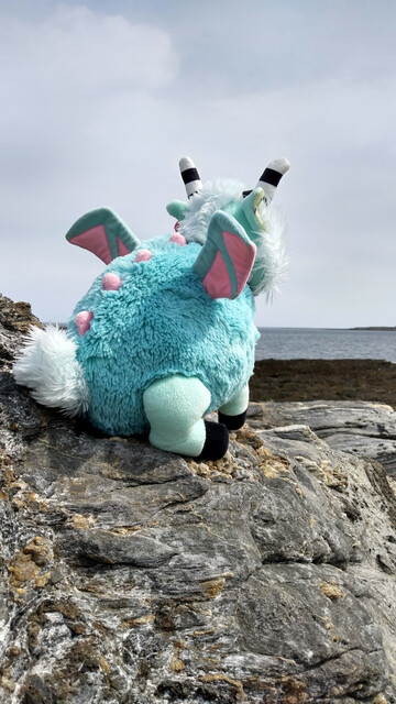 A pink and teal plushie demon, sitting on rocks by the ocean, with an island in the distance.