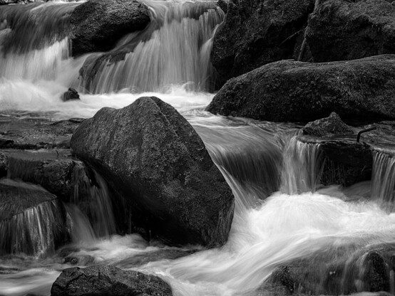 Mehrere Kaskadenstufen eines kleinen Bachwasserfalls mit bemoosten Steinen, an denen sich die Kaskaden entlangschlÃ¤ngeln.
Aufnahme wurde in SchwarzweiÃŸ und mit einer Langzeitbelichtung angefertigt.