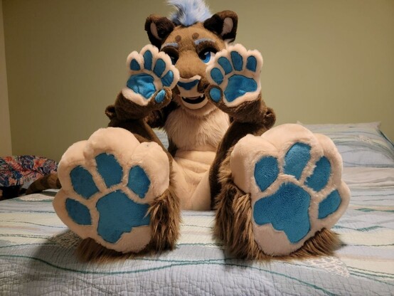 Lioness fursuiter sitting on bed showing toe beans and hand paws