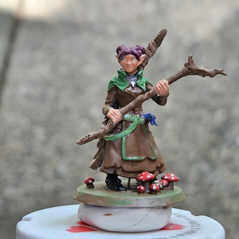 A dungeons and dragons mini of a circle of spores druid. She has purple hair, and green and brown robes. She has purple hair and green eyes. She is holding a staff and has a longbow on her back. There are mushrooms at her feet.