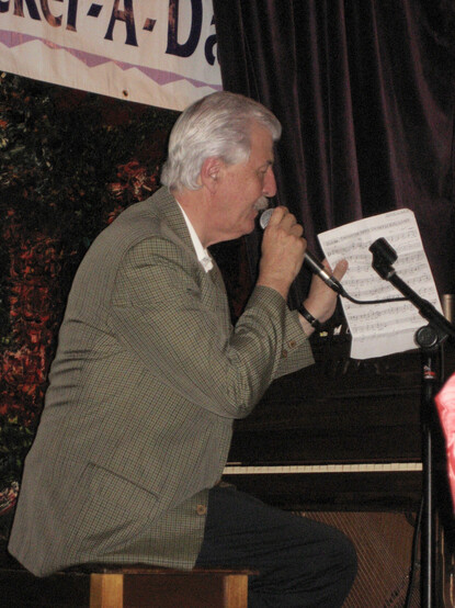 Man, pianist/composer/music historian Butch Thompson, sitting on stool of a piano.  He is wearing a suit, has grey hair and moustache. He holds a microphone in front of his mouth with his left hand. In his right hand he holds hand-written sheet music, with title visible "Swinging Down in New Orleans".