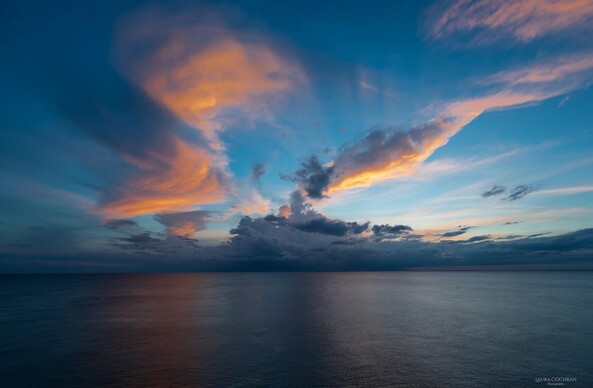 A wide angle photo of tall clouds reflecting the orange light, over dark blue water.