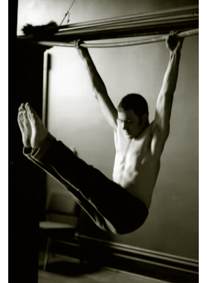 matt is a 22 year year-old athlete and actor, shirtless with jeans. he's improvising a gymnast's move. hanging from a pipe over a door way, legs up and level with his head, his eyes are closed. the setting is a doorway between two rooms in a rundown c. 1910 convent school.